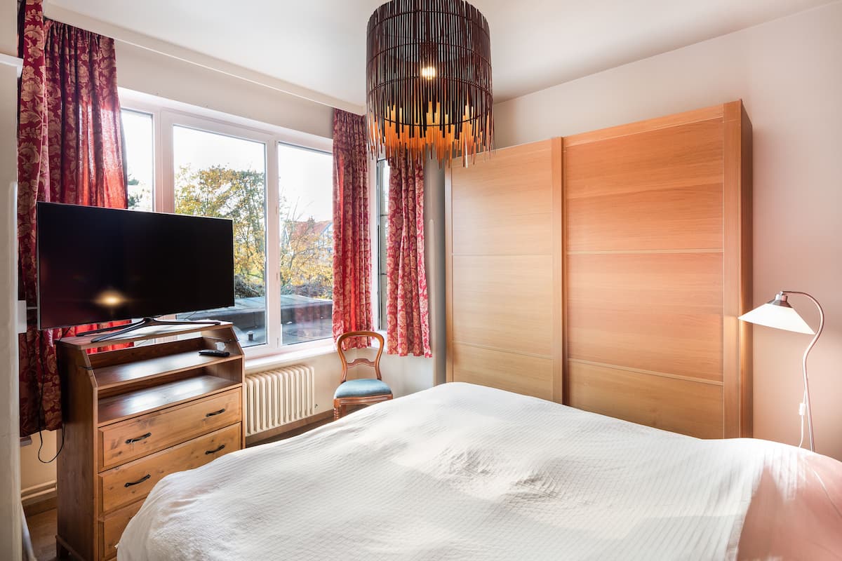 Ein Schlafzimmer mit einem Bett, einem Fernseher, einem Schminktisch, einem Stuhl, einem Kleiderschrank und einem Fenster mit Vorhängen.