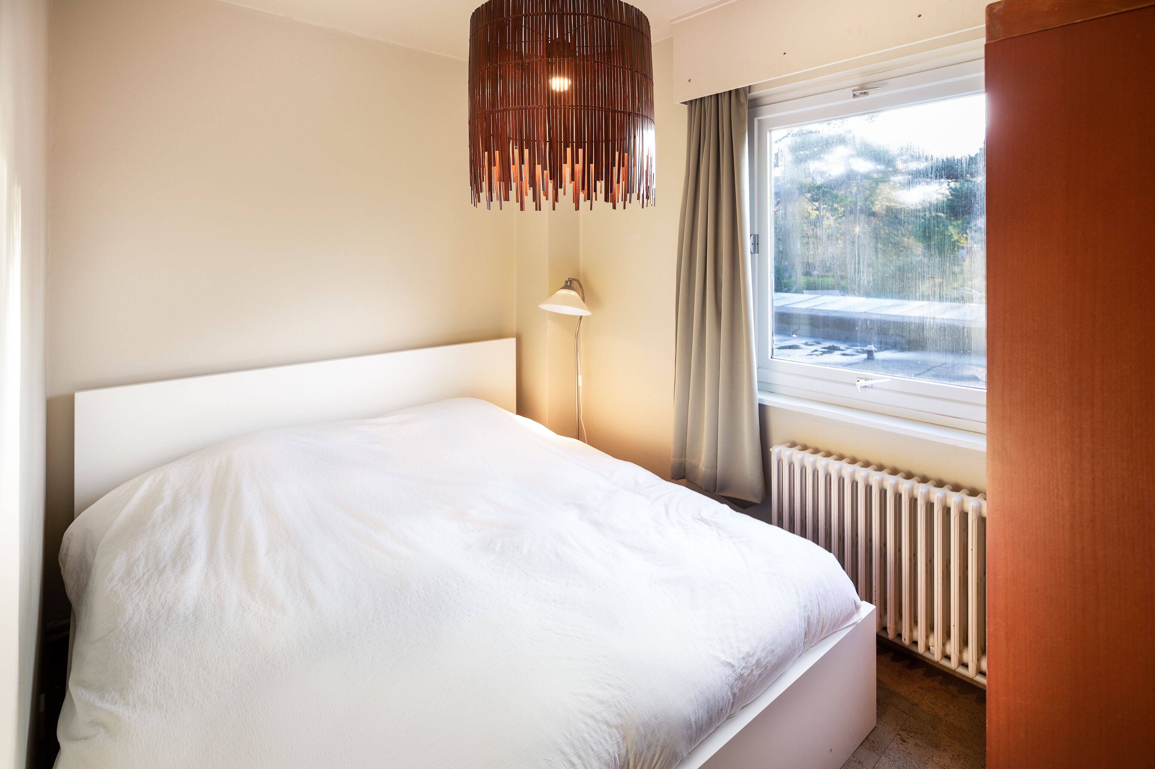 Une chambre à coucher avec un grand lit, une lampe sur pied, une fenêtre avec des rideaux et un radiateur.