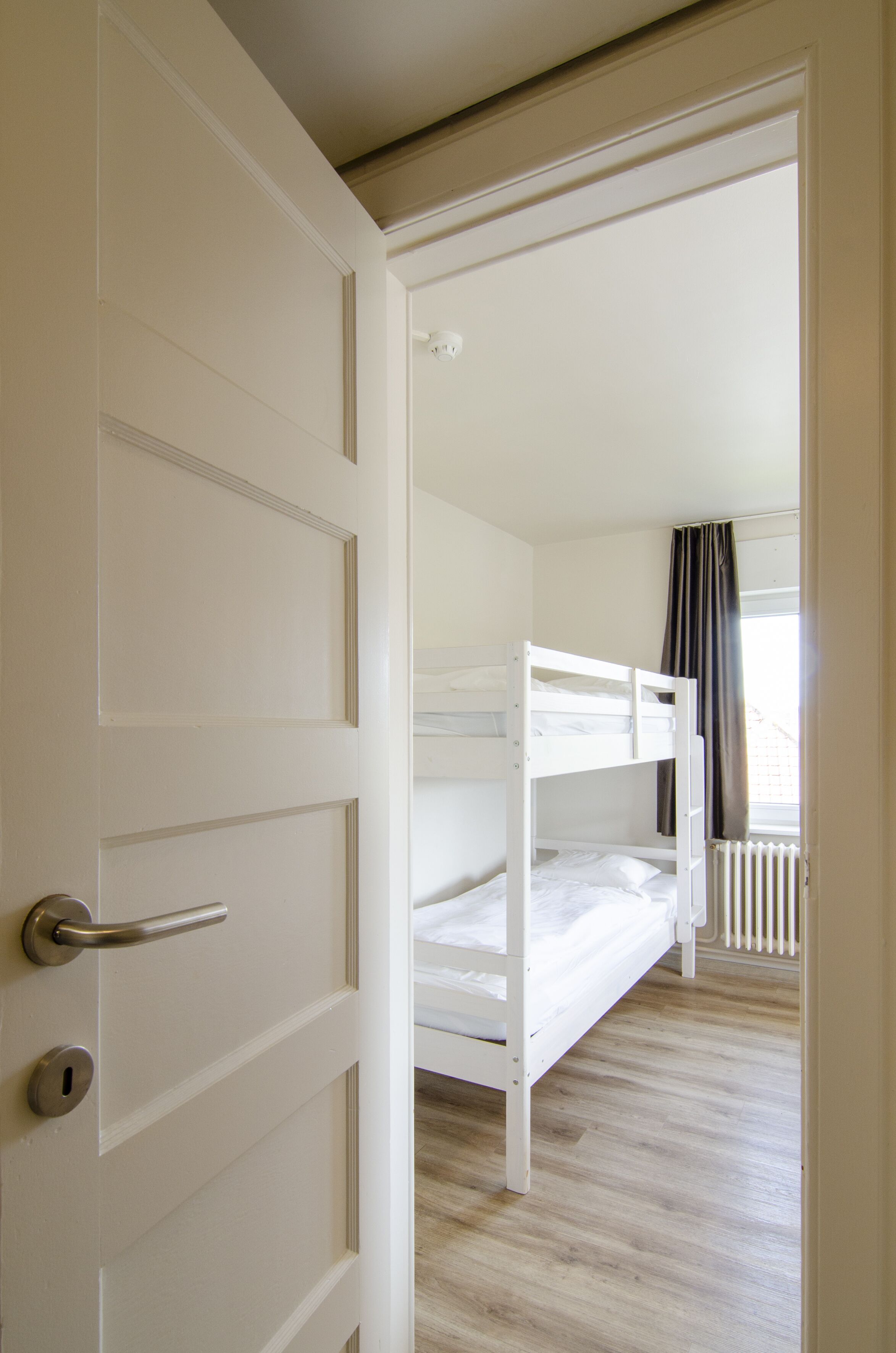 Una habitación con una cama litera, una puerta con manija y una ventana con cortinas.
