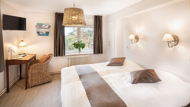 Une chambre d’hôtel avec un grand lit, un bureau avec une lampe, une chaise en osier, une fenêtre avec des rideaux et des lampes fixées au mur.