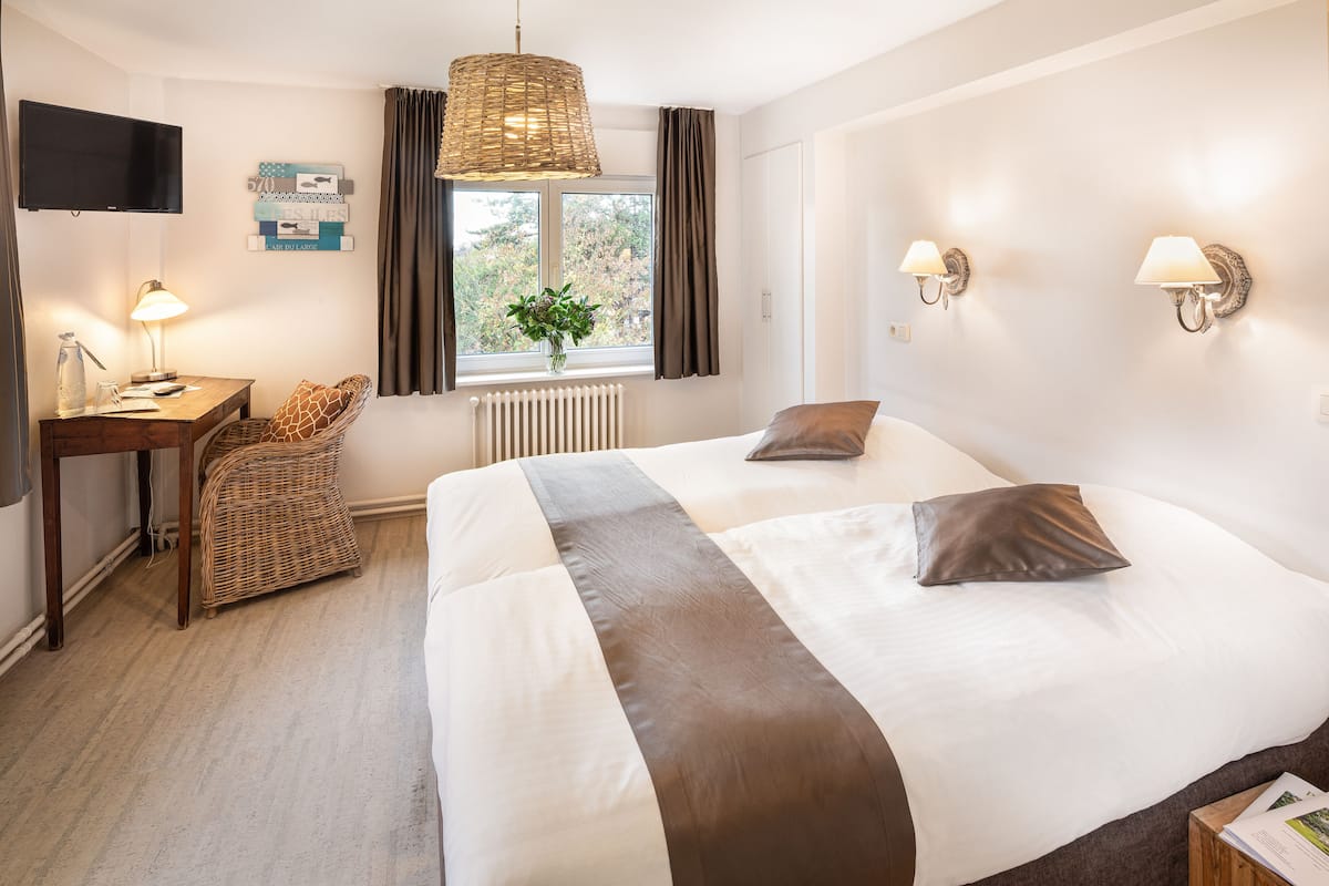 Ein Hotelzimmer mit einem großen Bett, einem Schreibtisch mit Lampe, einem Korbstuhl, einem Fenster mit Vorhängen und Wandlampen.