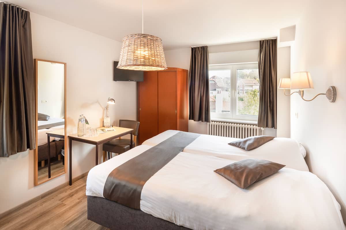 Ein Hotelzimmer mit zwei Betten, einem Schreibtisch, einem Stuhl, einem Spiegel, einem Fenster mit Vorhängen und einer Pendelleuchte.