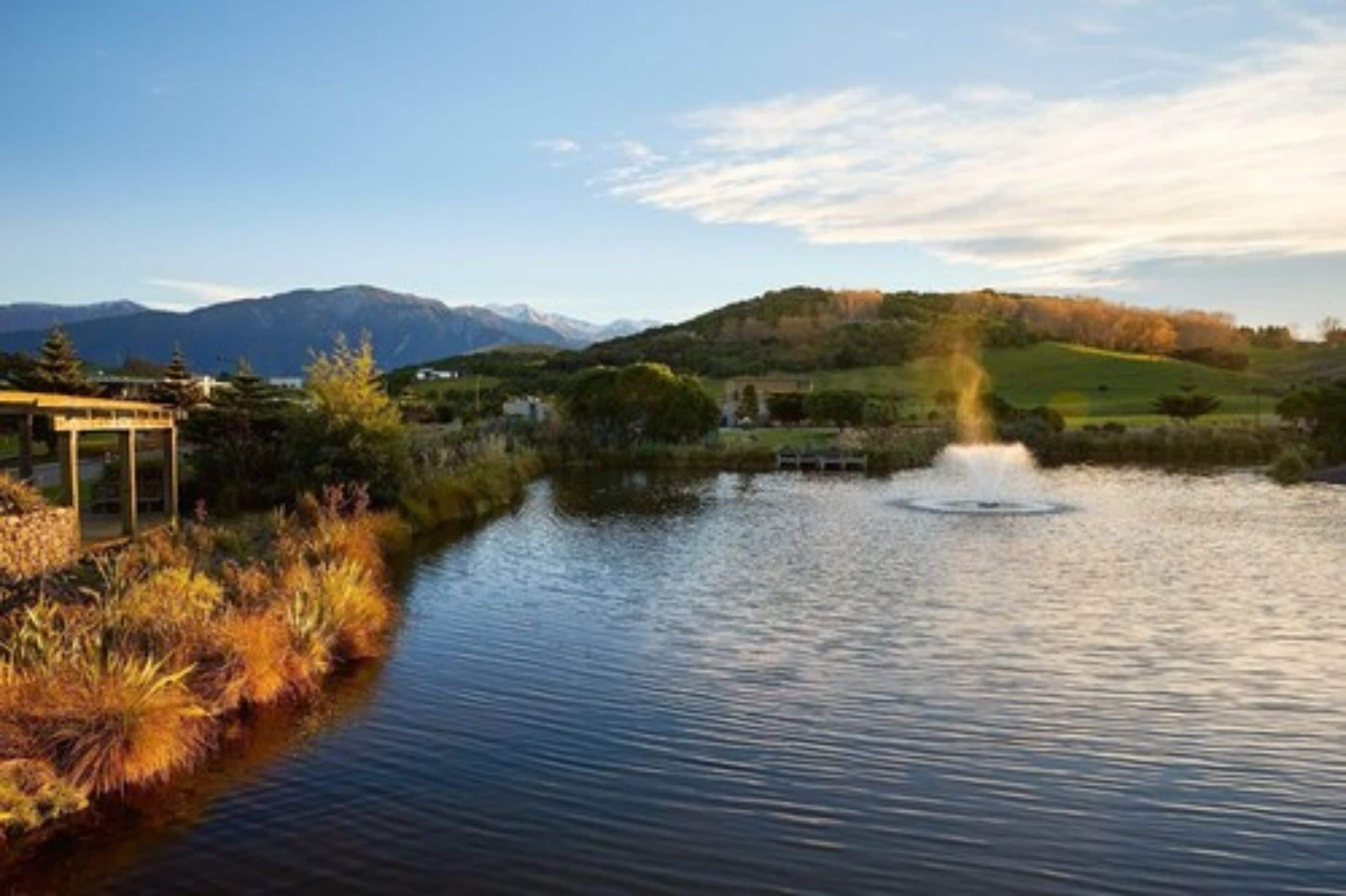 A serene lakeside view with a fountain, a building with a covered area, and mountains in the background.