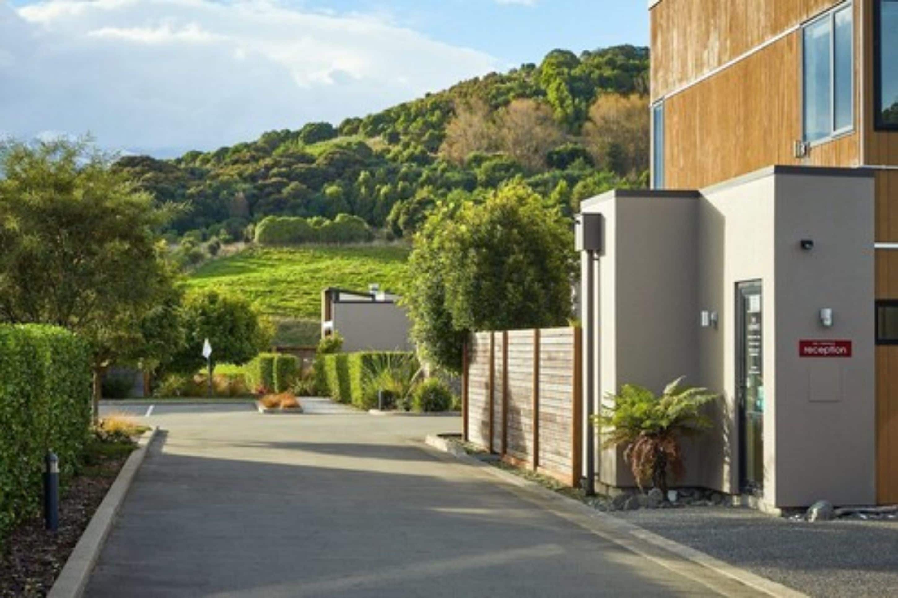 A modern building with a reception area, a paved driveway, and a hillside in the background.
