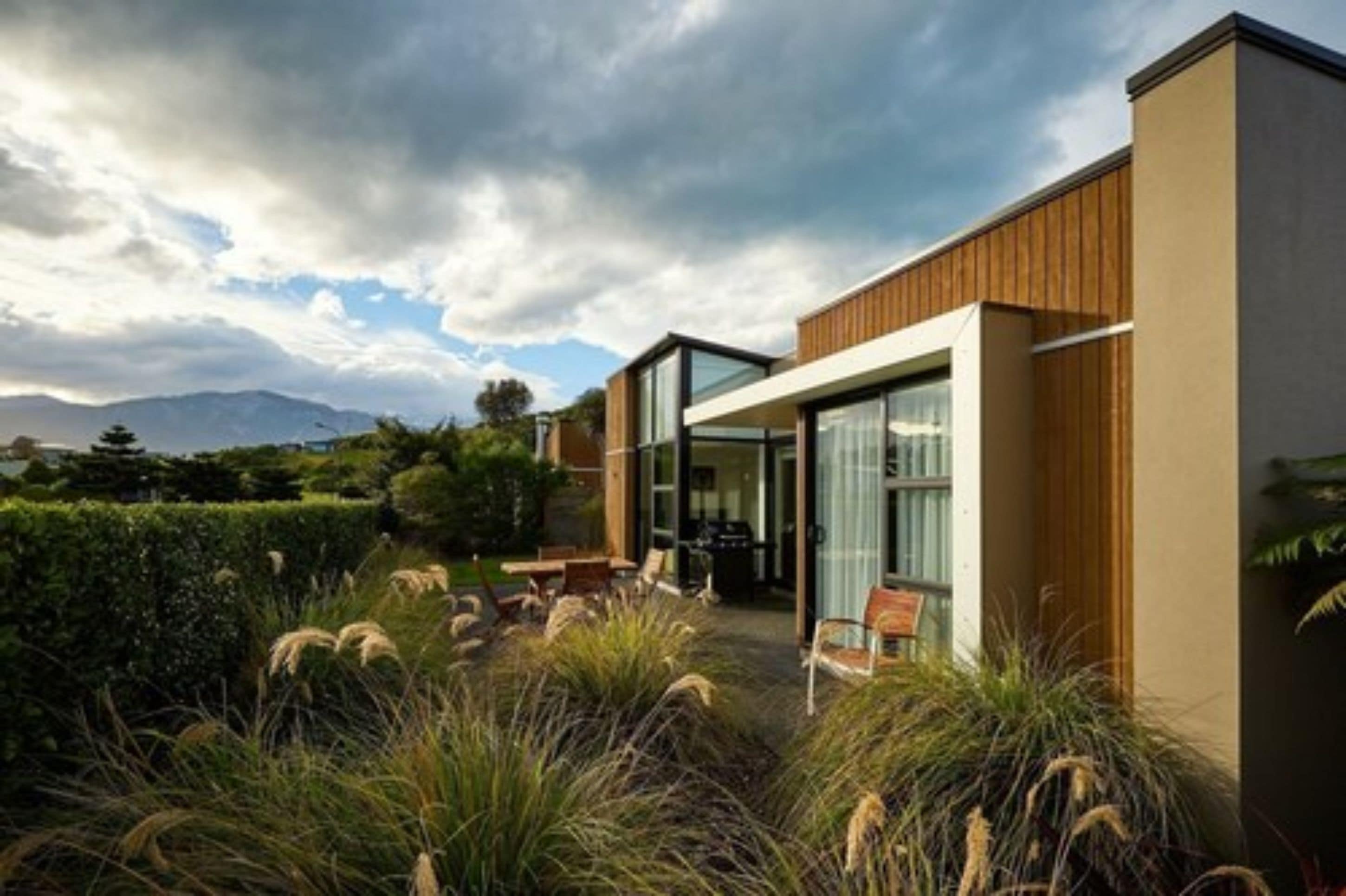 A modern house with a glass door entrance, surrounded by a well-maintained garden with tall grasses and a view of mountains in the background.