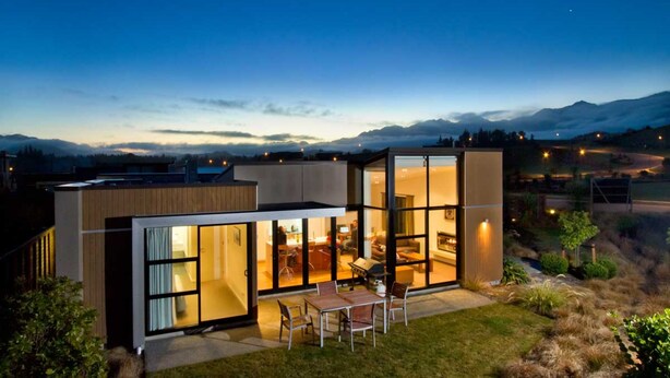 Une maison moderne avec une façade en verre, un espace salon extérieur et une vue sur les montagnes.