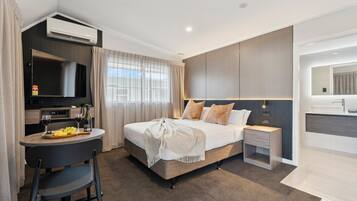 A modern hotel room with a bed, bedside table, TV, and a dining area with a table and chairs.