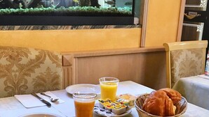 Una mesa de comedor dispuesta con un plato, cubiertos, un vaso de jugo de naranja, una cesta de croissants y un recipiente pequeño de mantequilla.