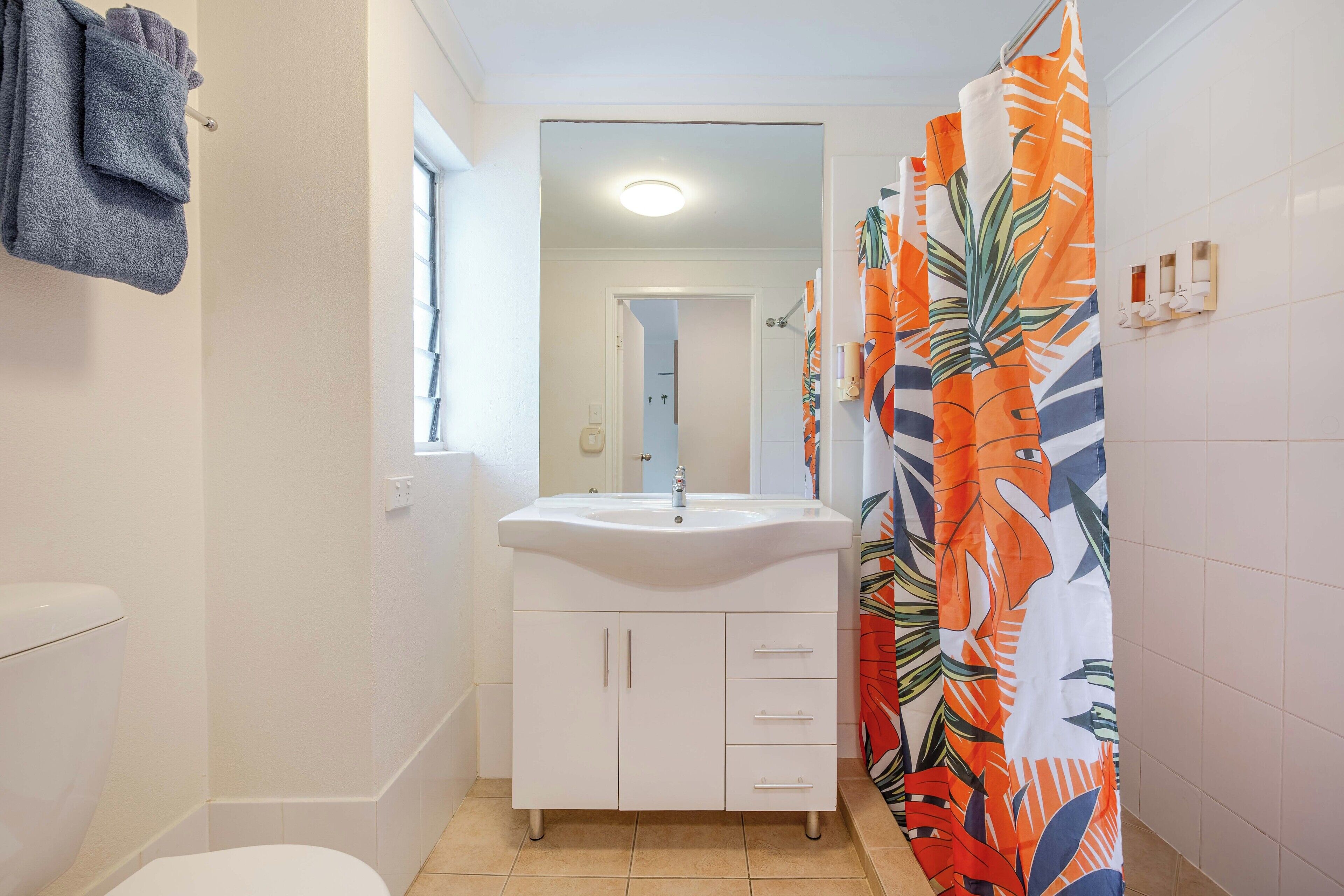 Un bagno con un lavandino bianco, un water e una tenda da doccia con motivo a foglia tropicale.