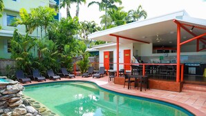 A swimming pool with lounge chairs and a covered bar area, surrounded by greenery and a building with red doors. - Port Douglas Motel (Port Douglas)