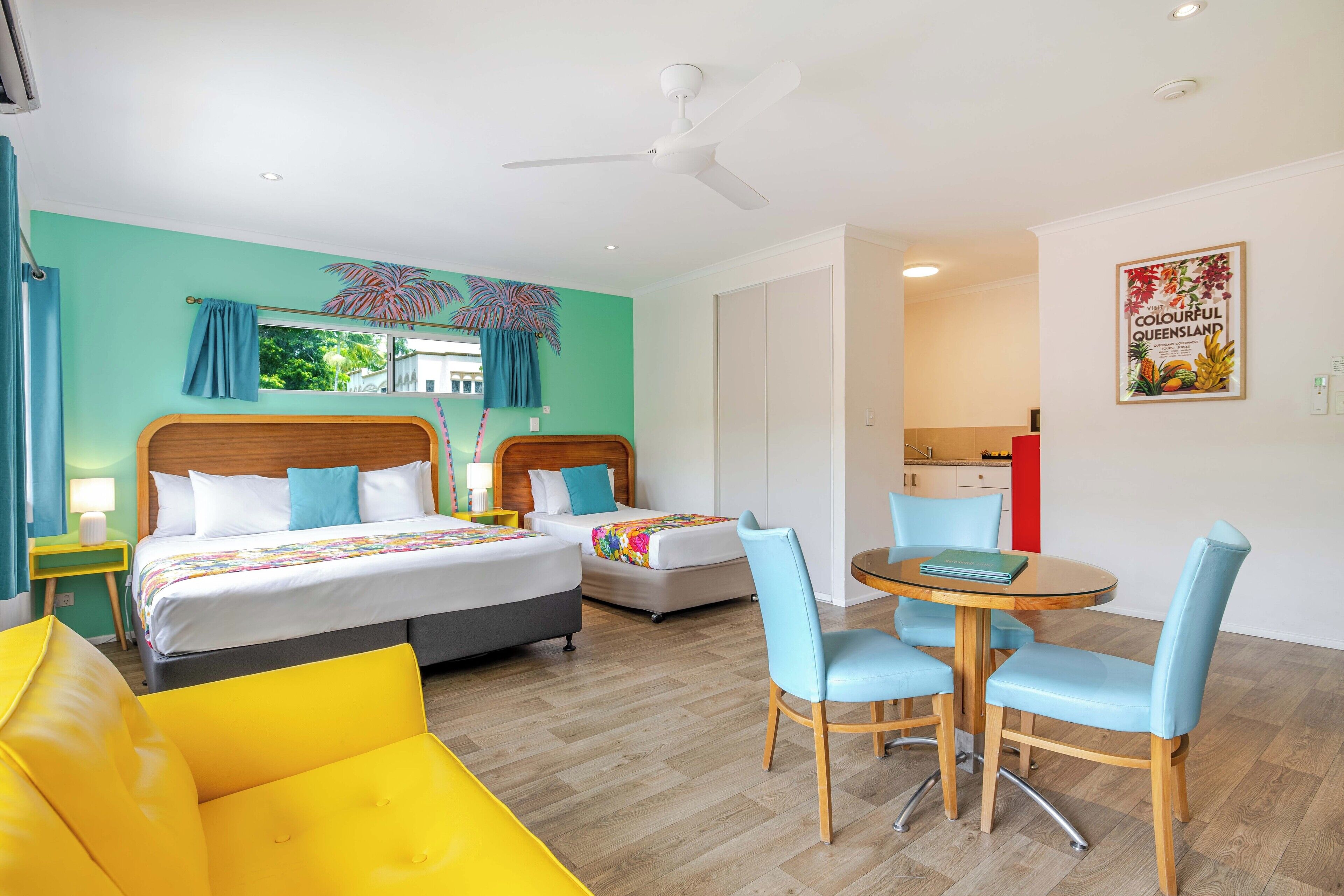 A hotel room with two beds, a dining table with chairs, a ceiling fan, and a kitchen area.