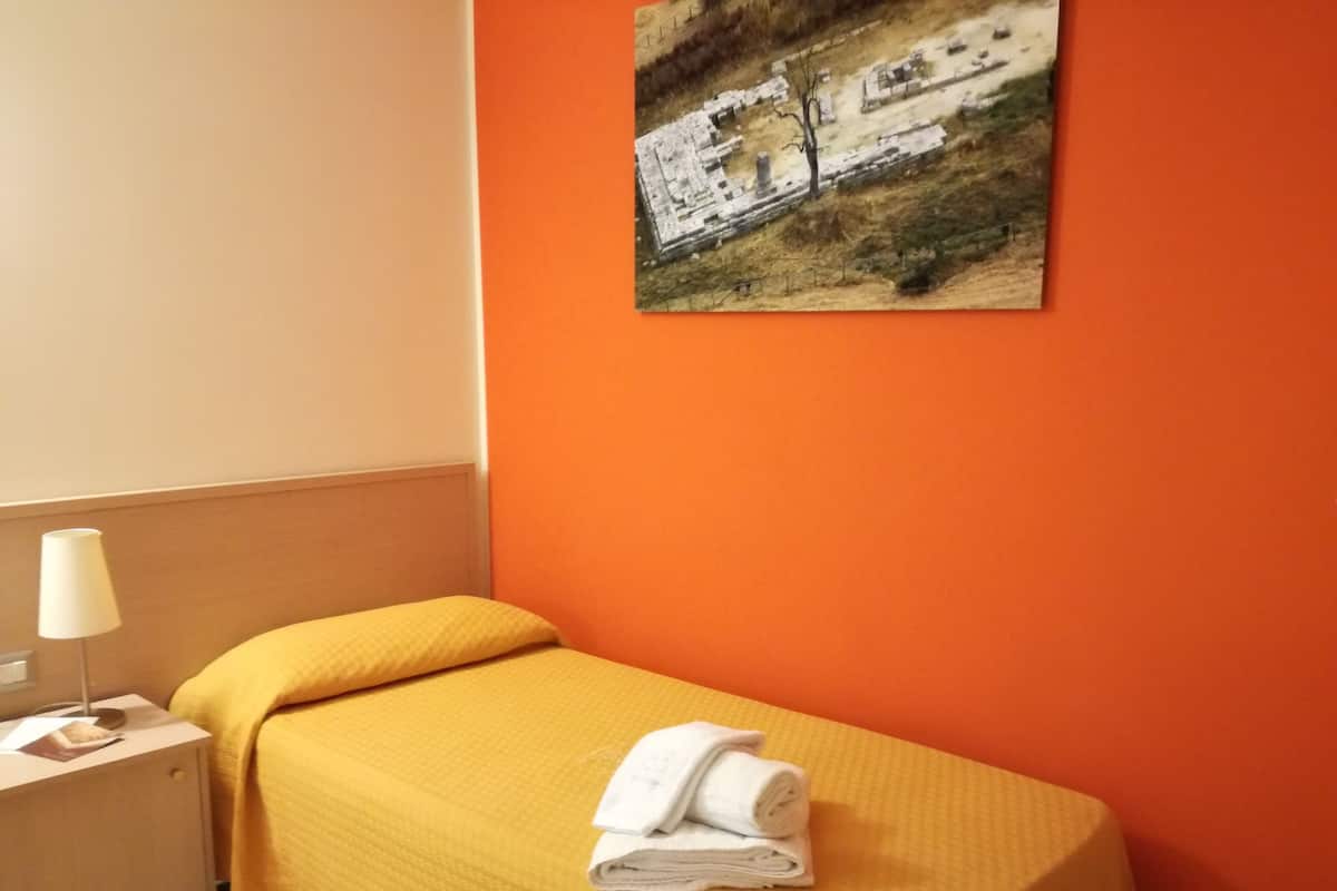 A single bed with a yellow bedspread and a rolled-up towel on it, a bedside table with a lamp, and a framed picture on the wall.
