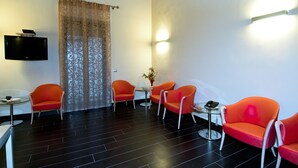 A hotel room with a dark tiled floor, orange chairs, a small table, a TV mounted on the wall, and a decorative curtain. - Hotel Residence Empedocle (Messina)