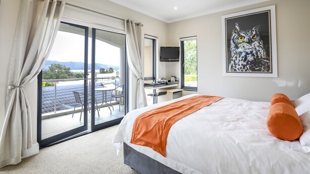 A bedroom with a large bed, an orange throw, a desk with a chair, a TV, and a balcony with a view. - Orange Inn Knysna (Knysna)