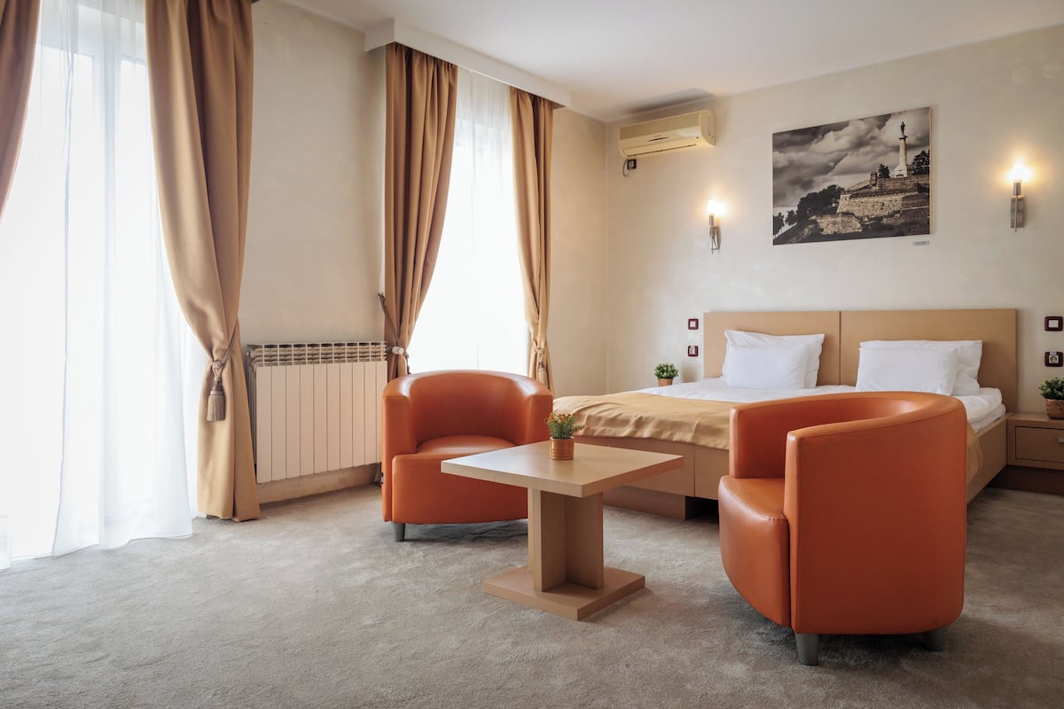 A hotel room with a bed, two orange armchairs, a small table, a radiator, a wall-mounted air conditioner, a painting, and a potted plant.