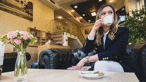 A woman in a navy blazer and white pants sitting on a black leather couch in a cafe, holding a white cup. There is a vase with pink flowers on the table, and a menu is visible. - Euro Garni Hotel (Belgrade)