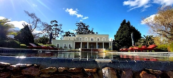 Um edifício grande de dois andares com fachada simétrica, entrada central e área de piscina ao redor.