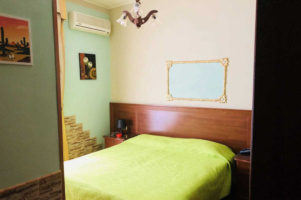 Ein Zimmer mit einem Bett, einem Nachttisch, einem Wandspiegel und einem Bild von Sonnenblumen.