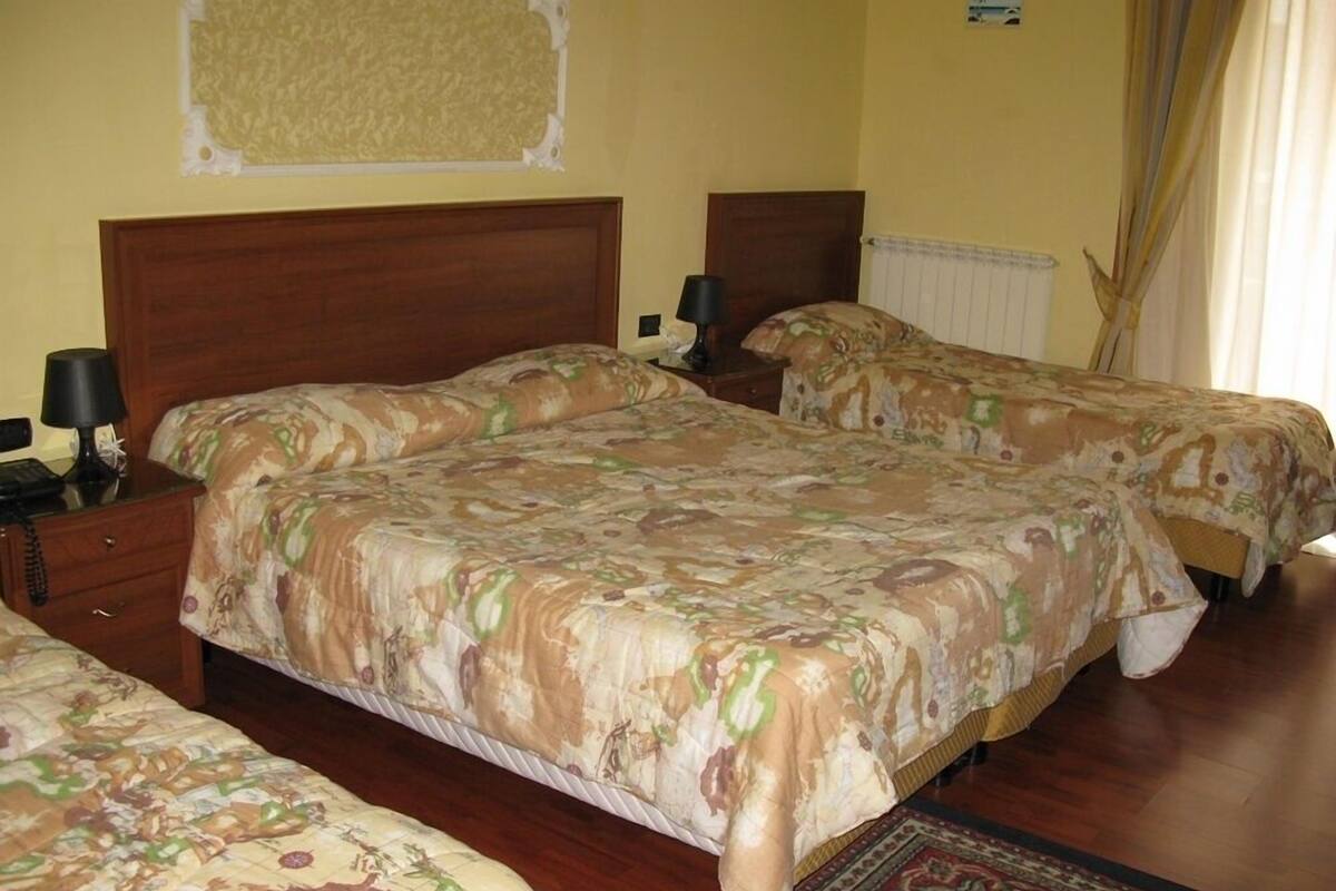 Ein Hotelzimmer mit einem großen Bett, zwei Einzelbetten, hölzernen Kopfteilen, Nachttischen mit Lampen, einem Telefon und einem gemusterten Teppich.