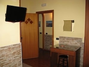 A room with a wooden door, a television mounted on the wall, a small table with a stool, and a stone-patterned wall. - Hotel Iris (Naples)