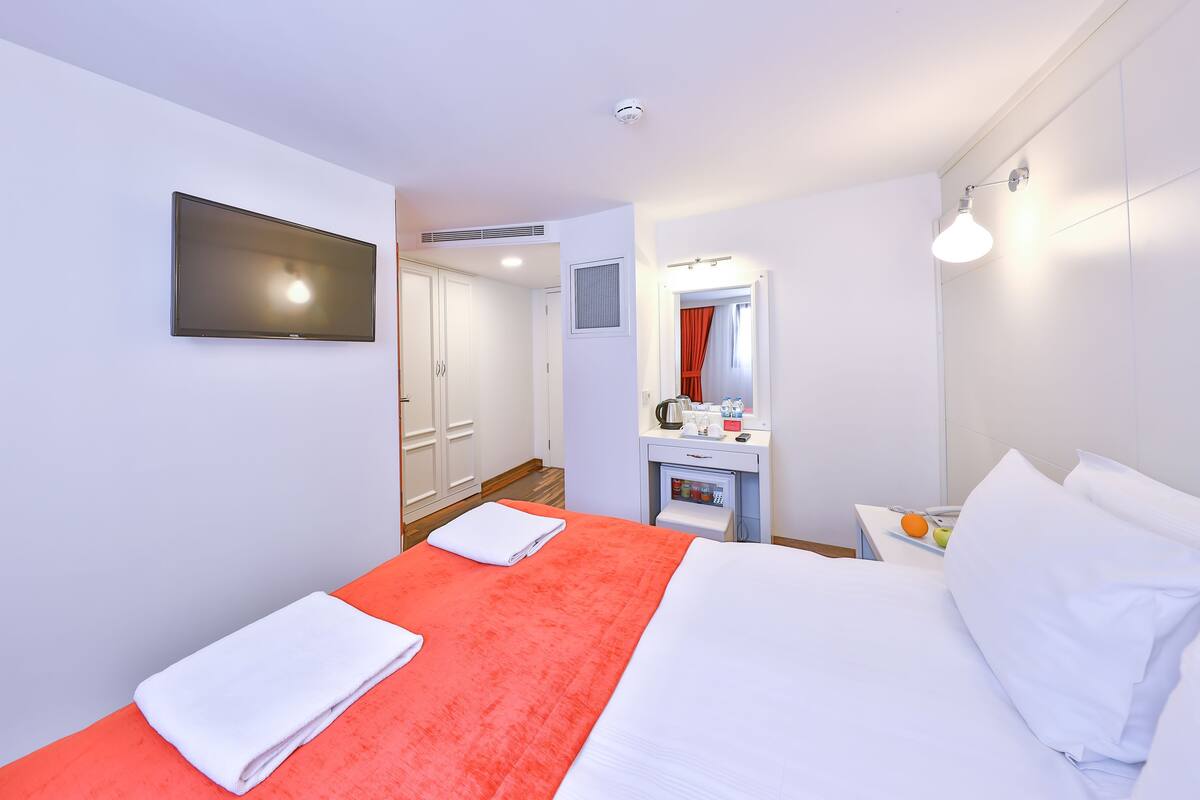 A hotel room with a bed, a TV mounted on the wall, a small table with a kettle, and a mirror.