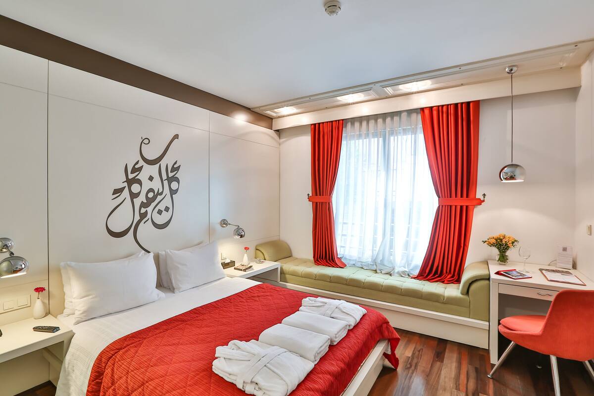 A modern hotel room with a bed, a desk, a chair, a window with red curtains, and a wall with decorative script.