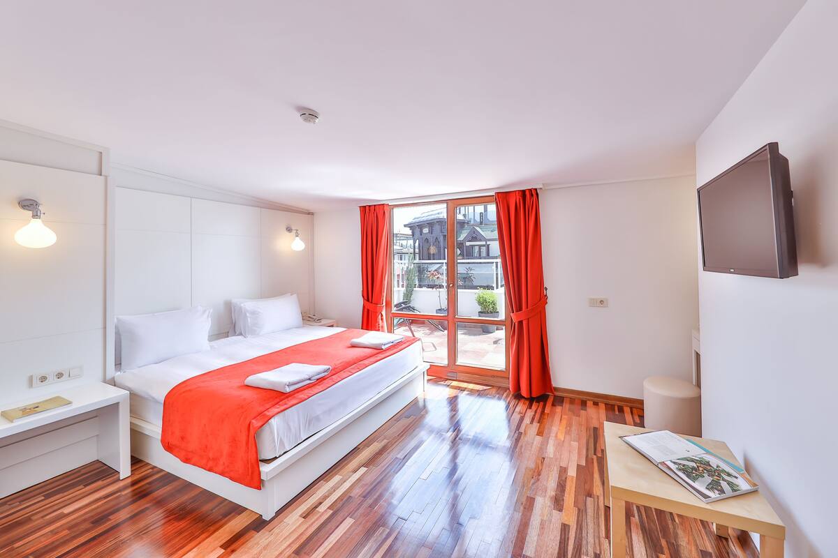A modern hotel room with a large bed, a small desk, a TV, and a balcony with red curtains.