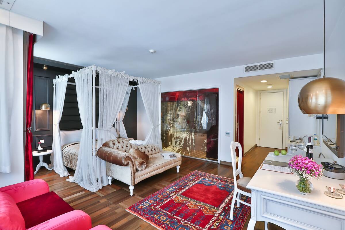A bedroom with a four-poster bed, a sitting area with a sofa, a desk with a lamp, and a red rug.