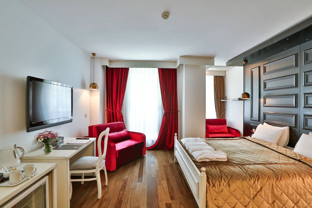 A hotel room with a bed, a desk with a chair, a TV, and a window with red curtains.
