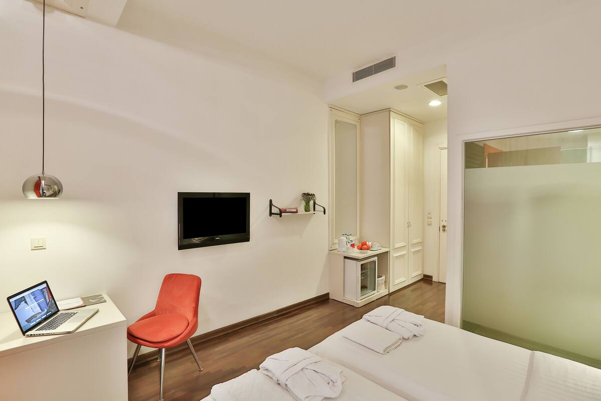 A hotel room with a desk, a laptop, a TV, a red chair, two beds, a mirror, and a wardrobe.