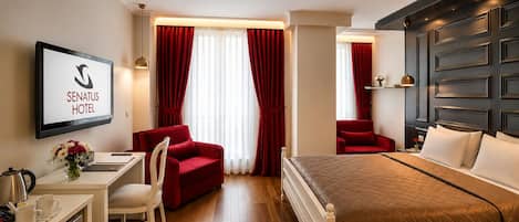 Habitación de hotel con una cama, dos sillas rojas, un escritorio con una computadora y un televisor mostrando 'SENATUS HOTEL'.