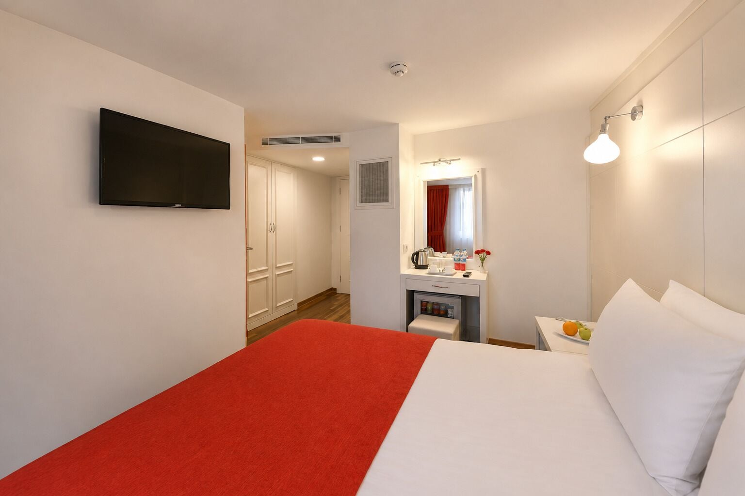 A hotel room with a bed, a TV mounted on the wall, a desk with a mirror, and a small kitchenette area.