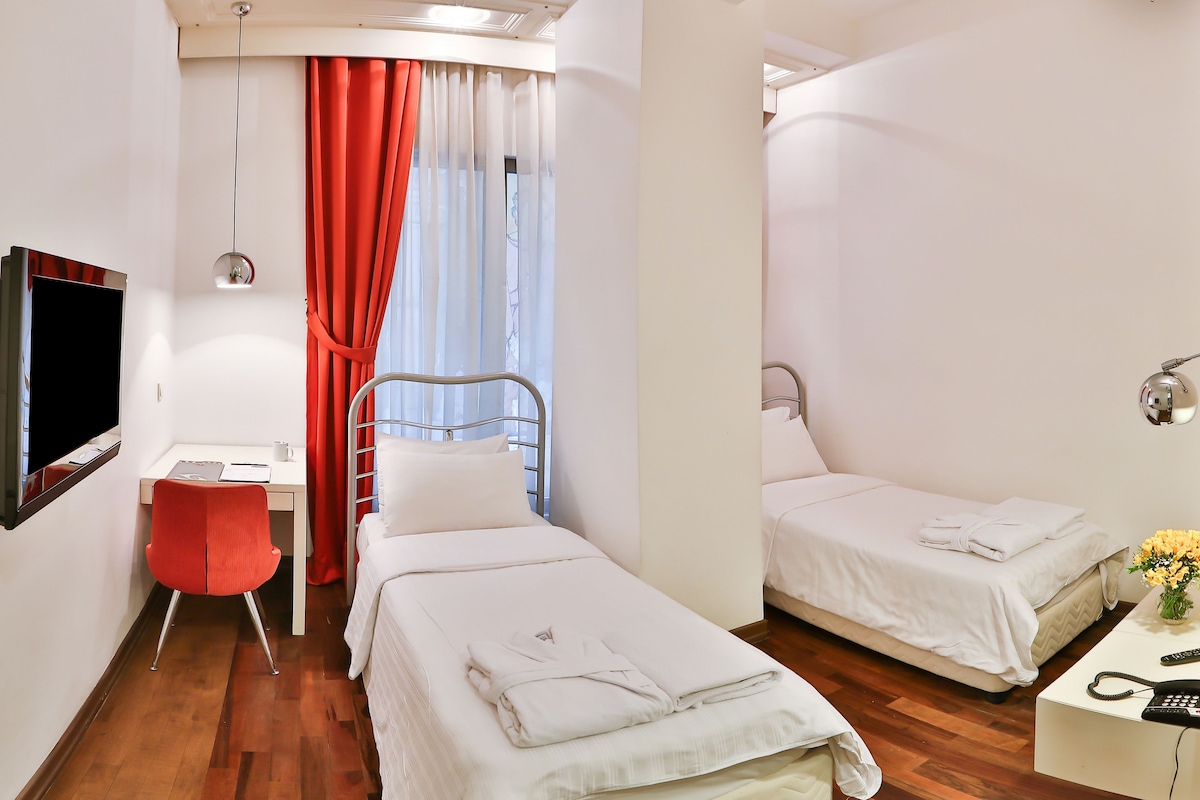 Quarto de hotel com cama, escrivaninha, cadeira, televisão e janela com cortinas.