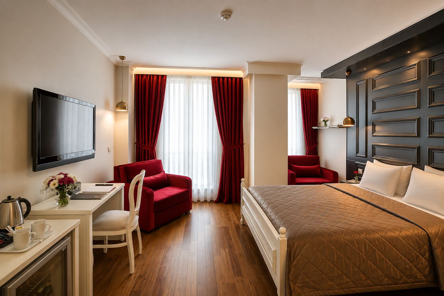 A hotel room with a bed, two red armchairs, a desk with a TV, and a minibar.
