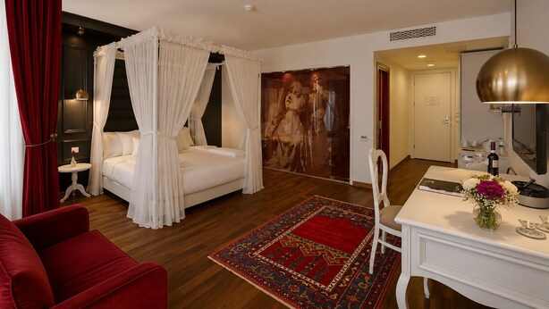 A hotel room with a four-poster bed, a red armchair, a desk with a mirror, and a large painting on the wall. - Senatus Hotel - Special Class (Istanbul)