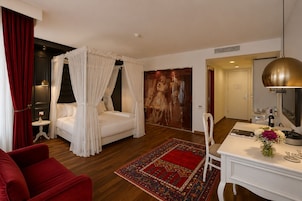 Habitación de hotel con cama con dosel, un sillón rojo, un escritorio con espejo y un cuadro grande en la pared.