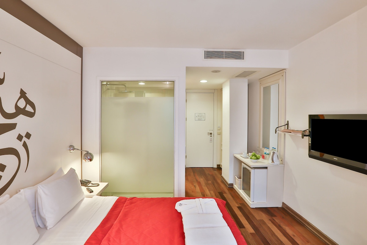 Ein Hotelzimmer mit Bett, roter Decke, Fernseher, Schreibtisch mit Spiegel und einer Tür zu einem Badezimmer.