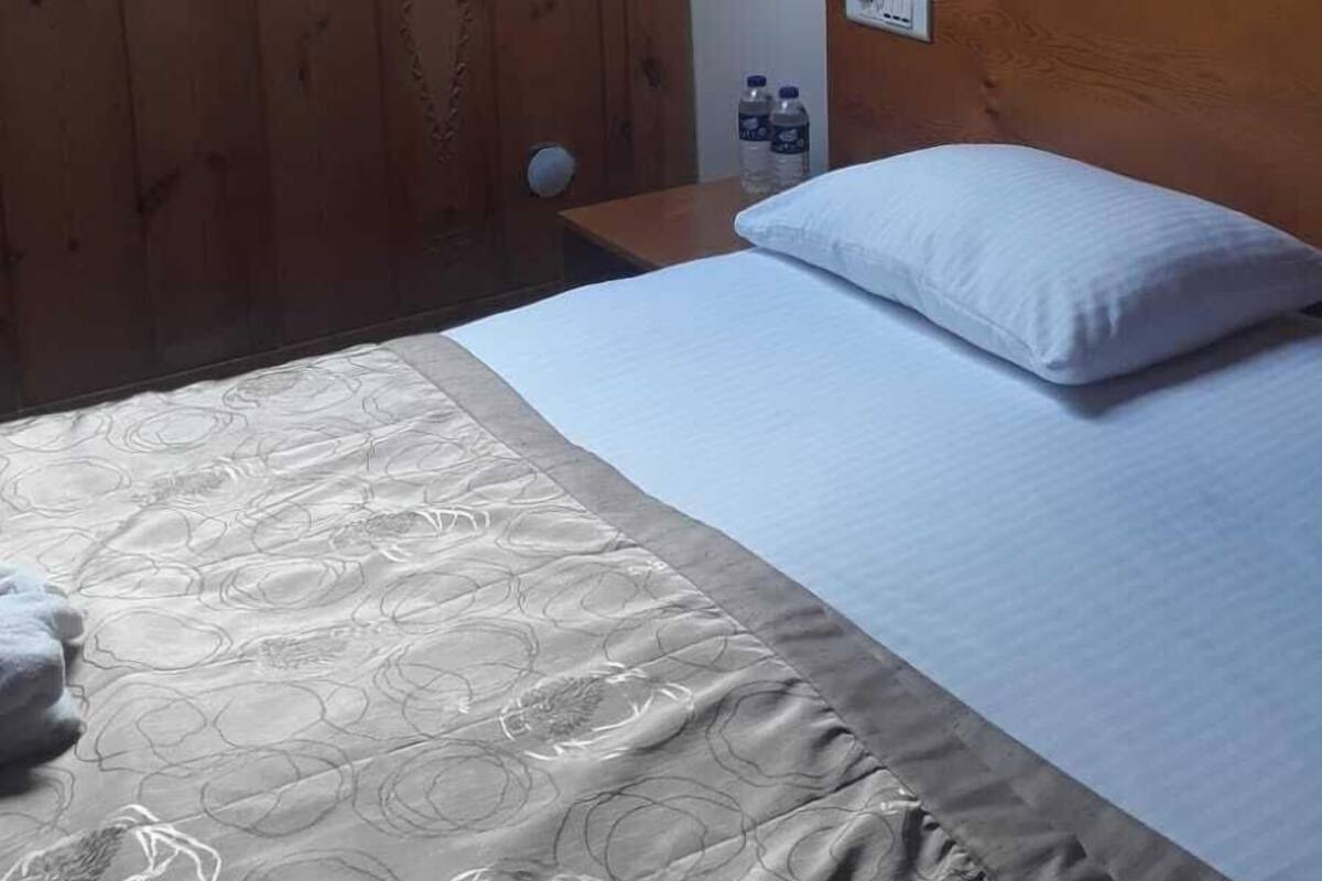 A room with a sloped ceiling, wooden walls, a bed with a white mattress and a grey headboard, a small bedside table with a lamp, and a skylight.