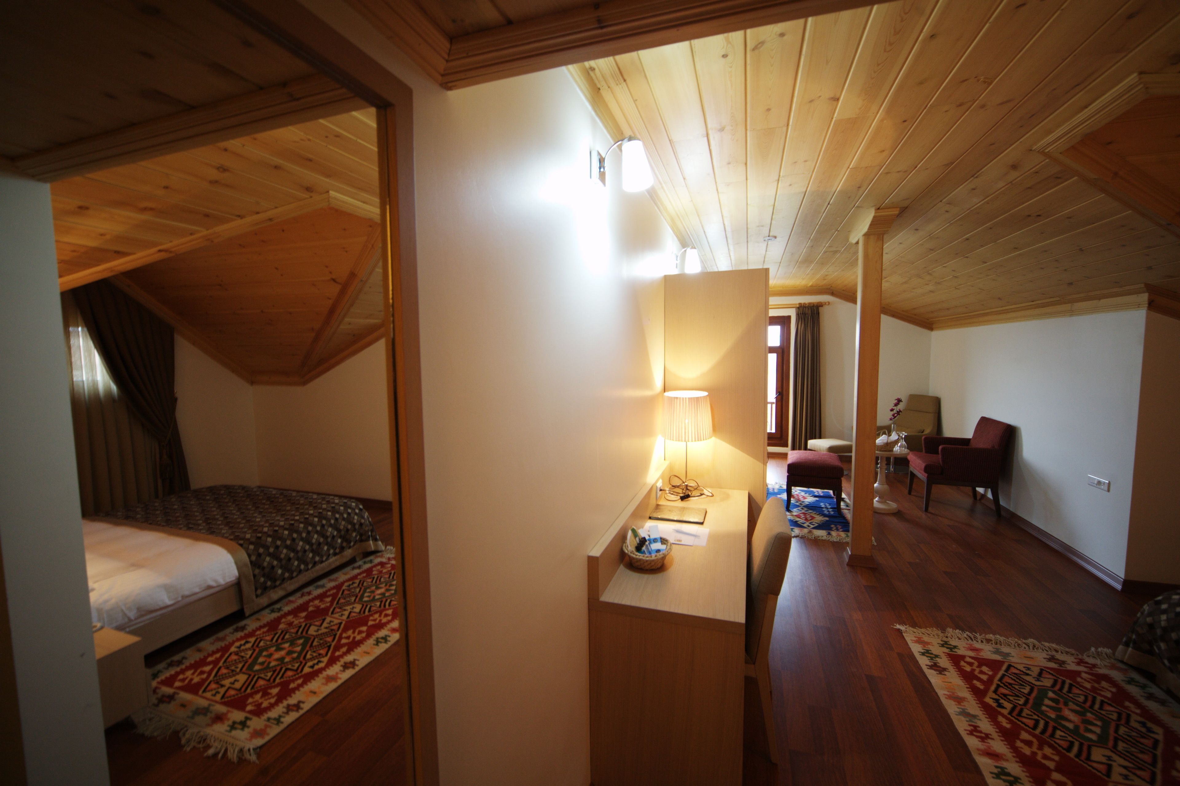 A room with a wooden ceiling, a bed with a patterned bedspread, a desk with a lamp, and a chair.