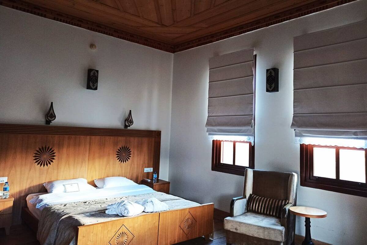 A bedroom with a wooden bed, a chair, a small table, and two windows with blinds.