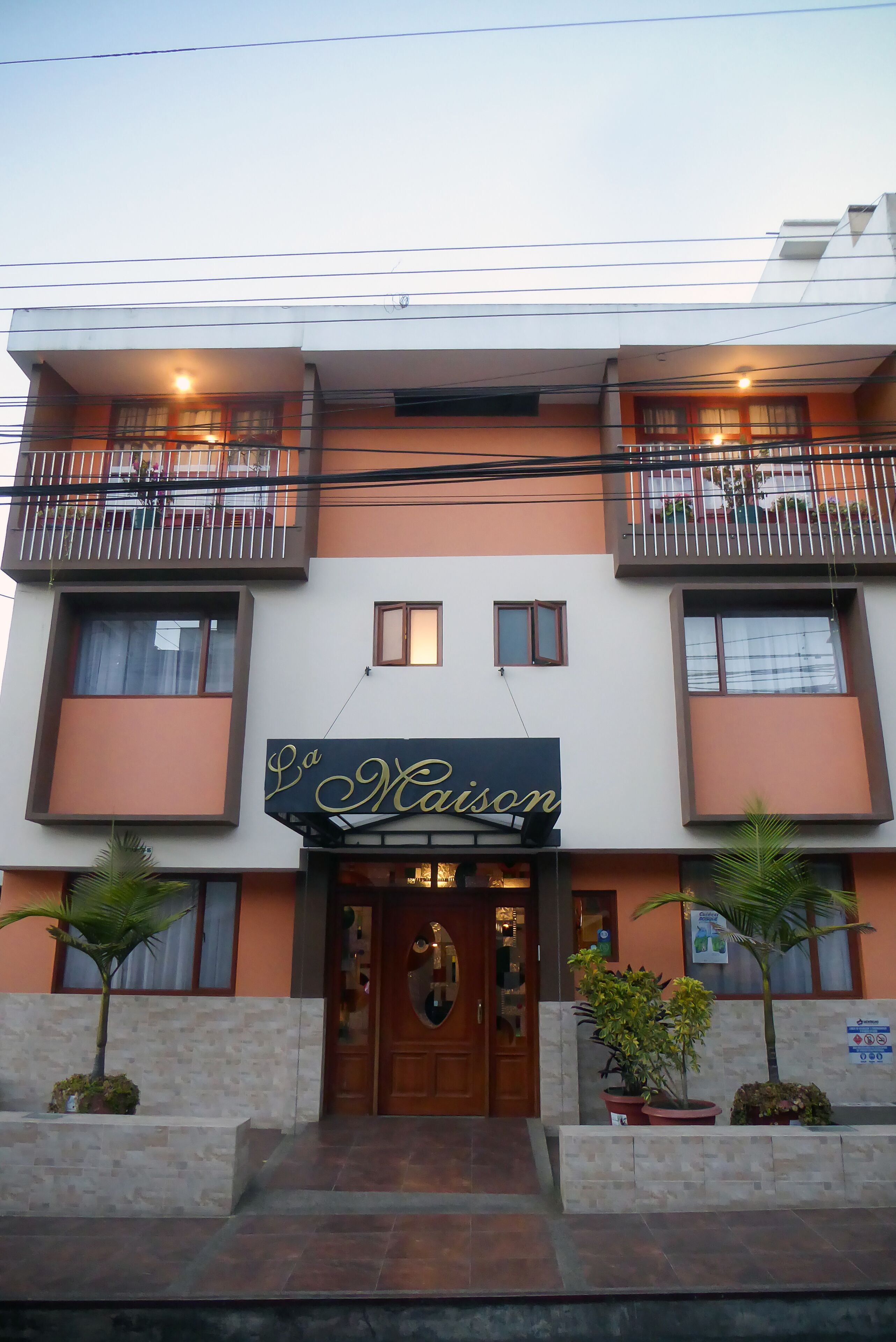A two-story building with a sign reading "La Maison" above the entrance. There are two balconies with railings, and the building has a modern architectural style.
