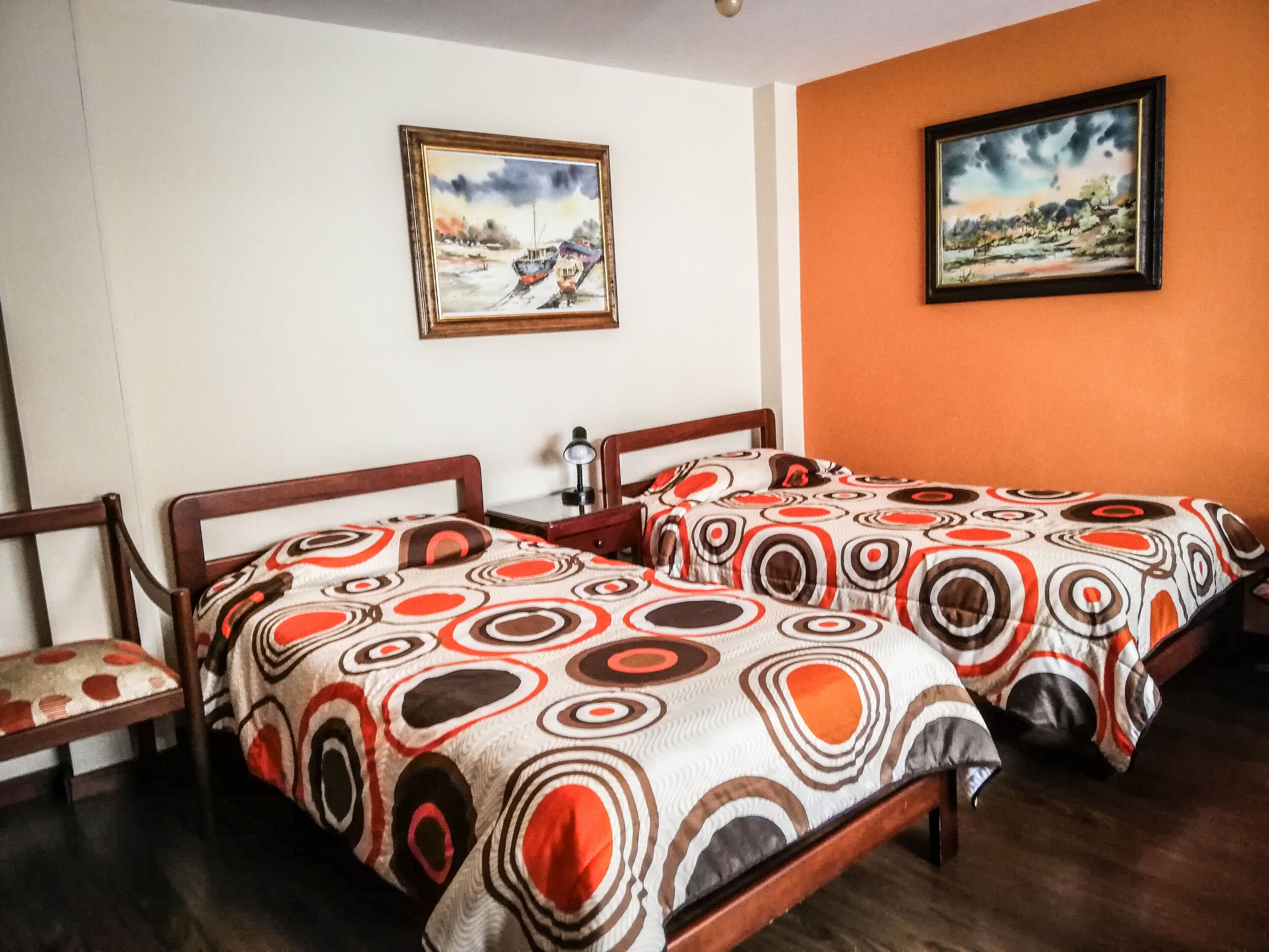 A hotel room with two wooden beds, a nightstand, a lamp, and two paintings on the walls.