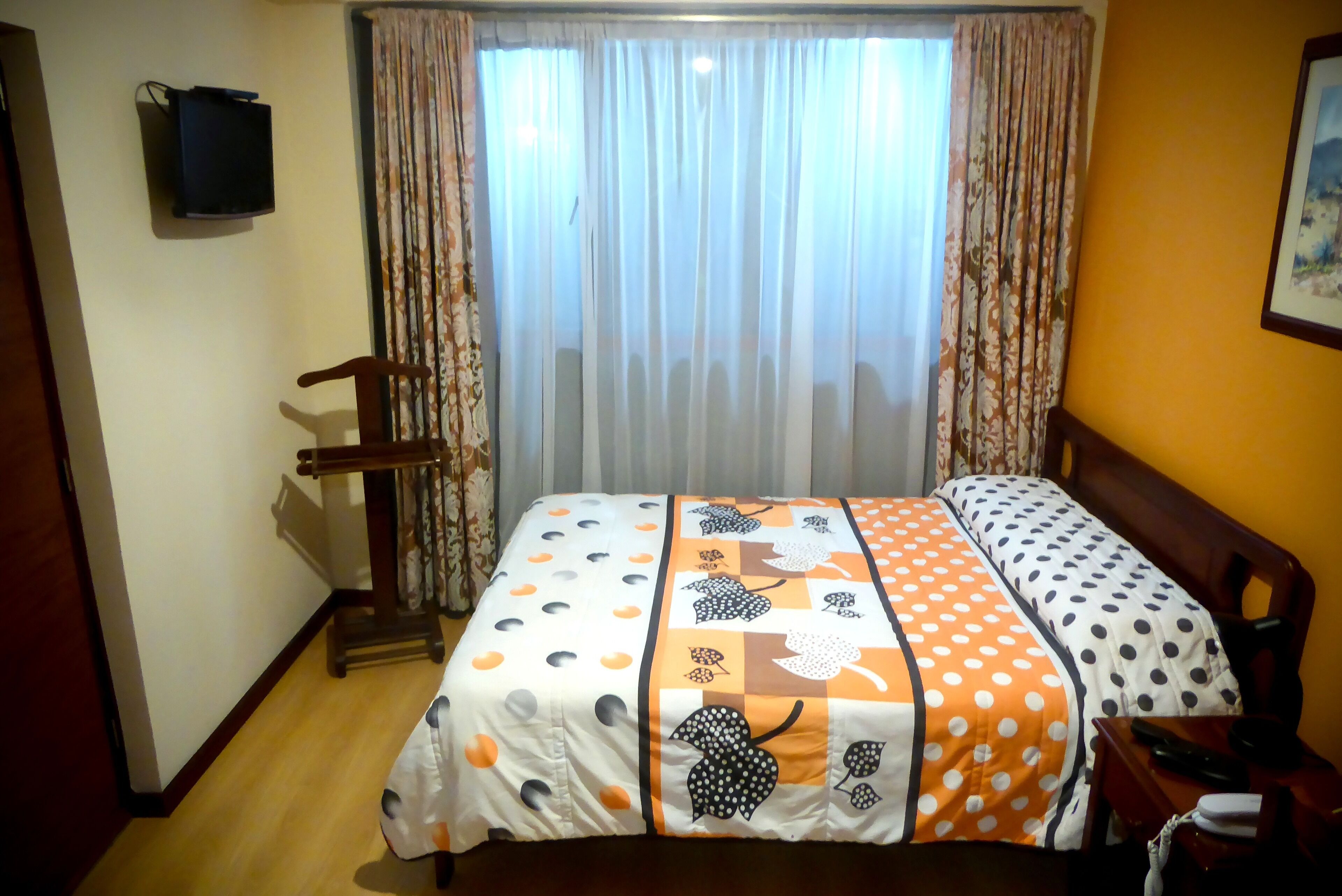 A neatly made bed with a patterned bedspread, a wooden chair, a small table with a phone, and a television on the wall.