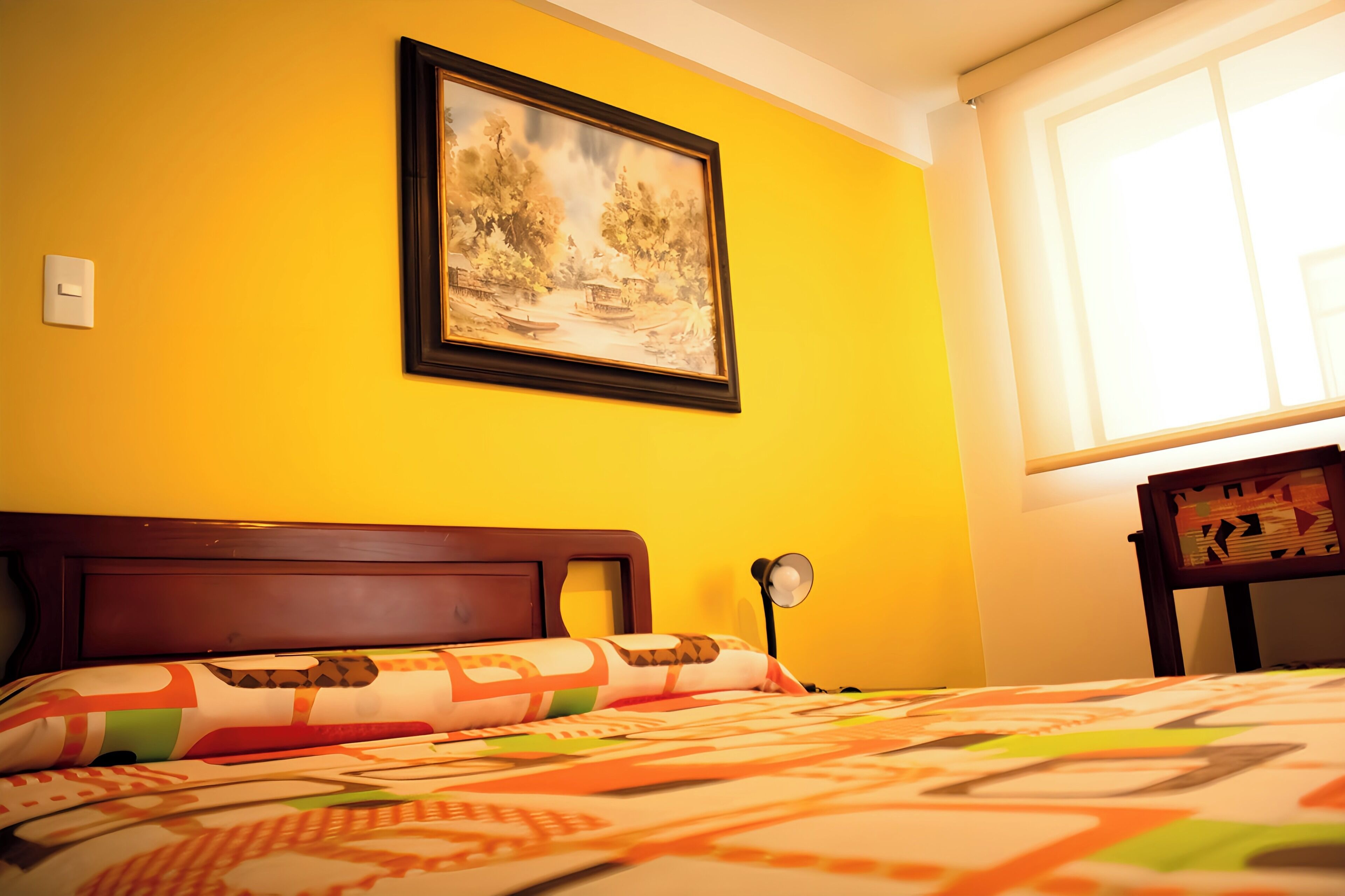 Una habitación con una cama, un cuadro enmarcado en la pared, una lámpara de noche y una ventana con luz natural.