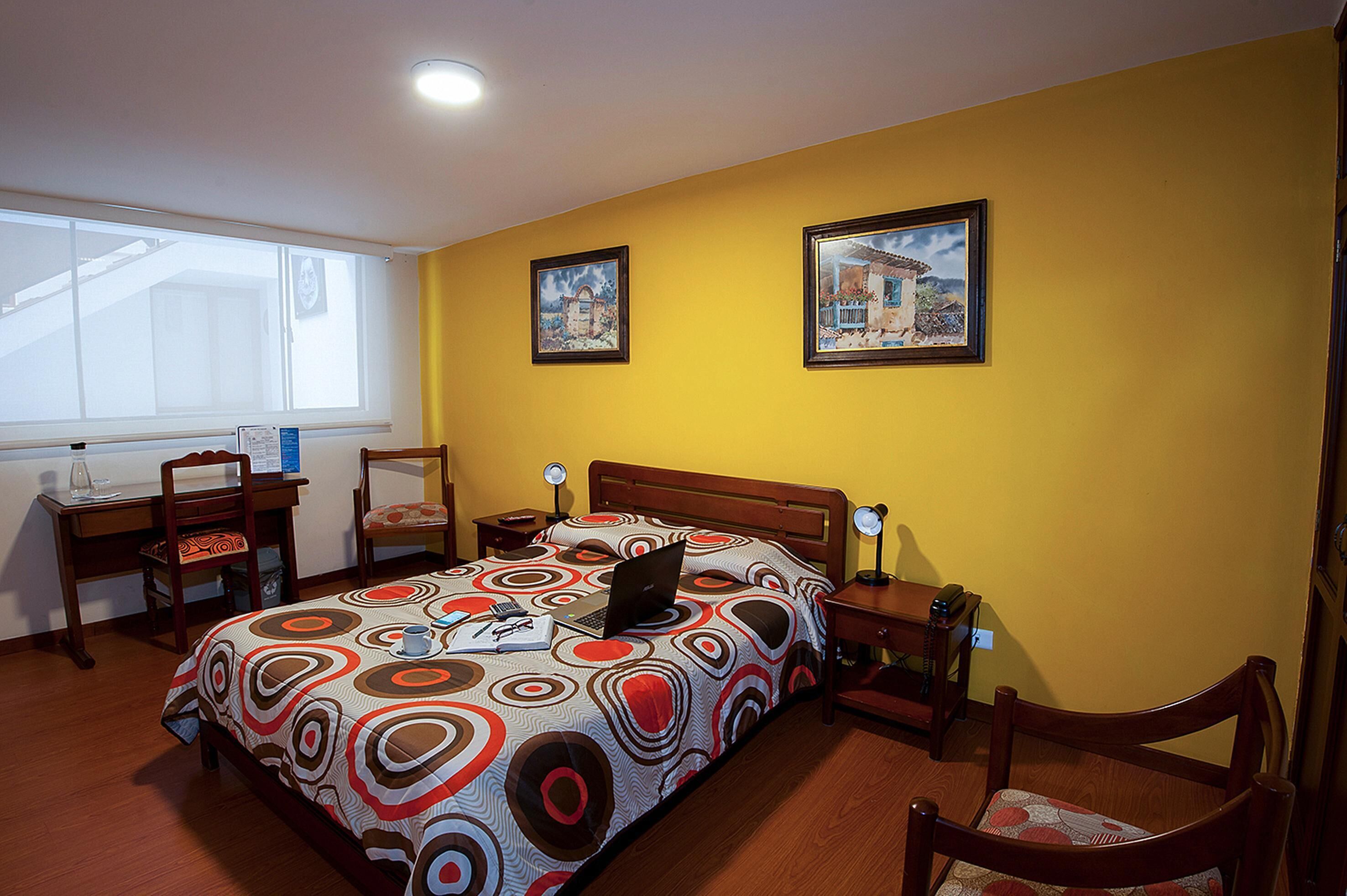 A bedroom with a bed, a desk, a chair, a nightstand, and two framed pictures on the wall.
