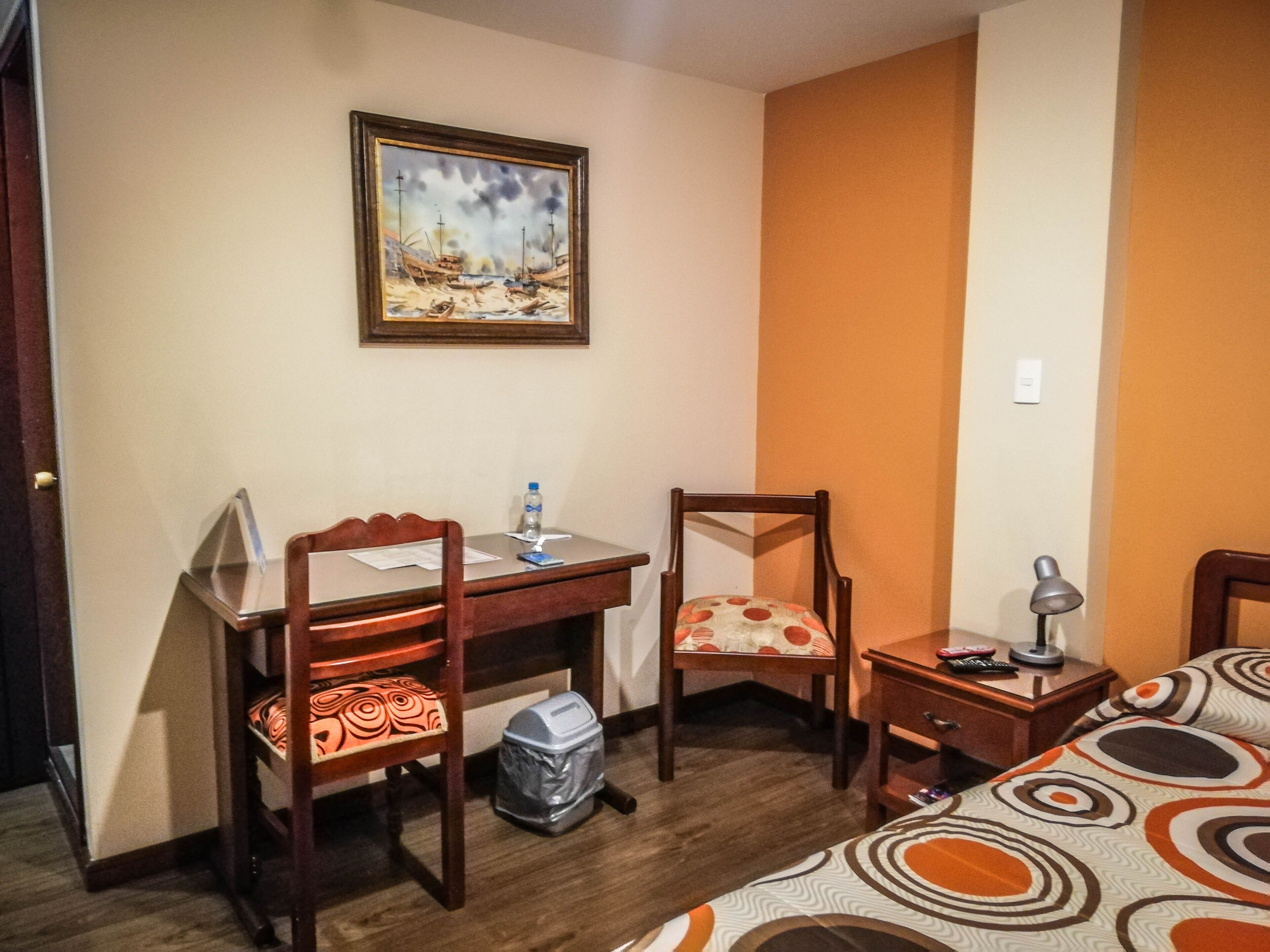 Habitación de hotel con un escritorio de madera, dos sillas, una cama con ropa de cama estampada, una mesita de noche con lámpara y un cuadro en la pared.