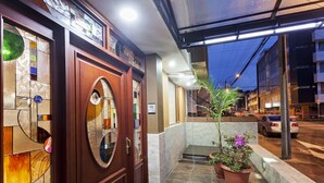 Een hotelentree met een houten deur, een glazen raam met kleurrijke patronen en een potplant op de stoep.