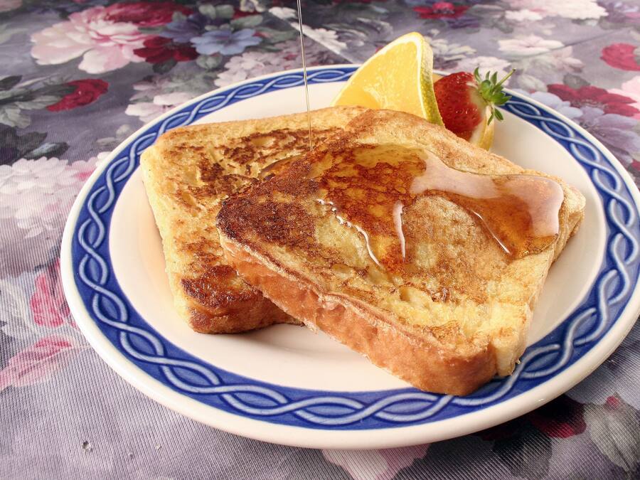 A plate with French toast, syrup, a lemon wedge, and a strawberry on a floral tablecloth.