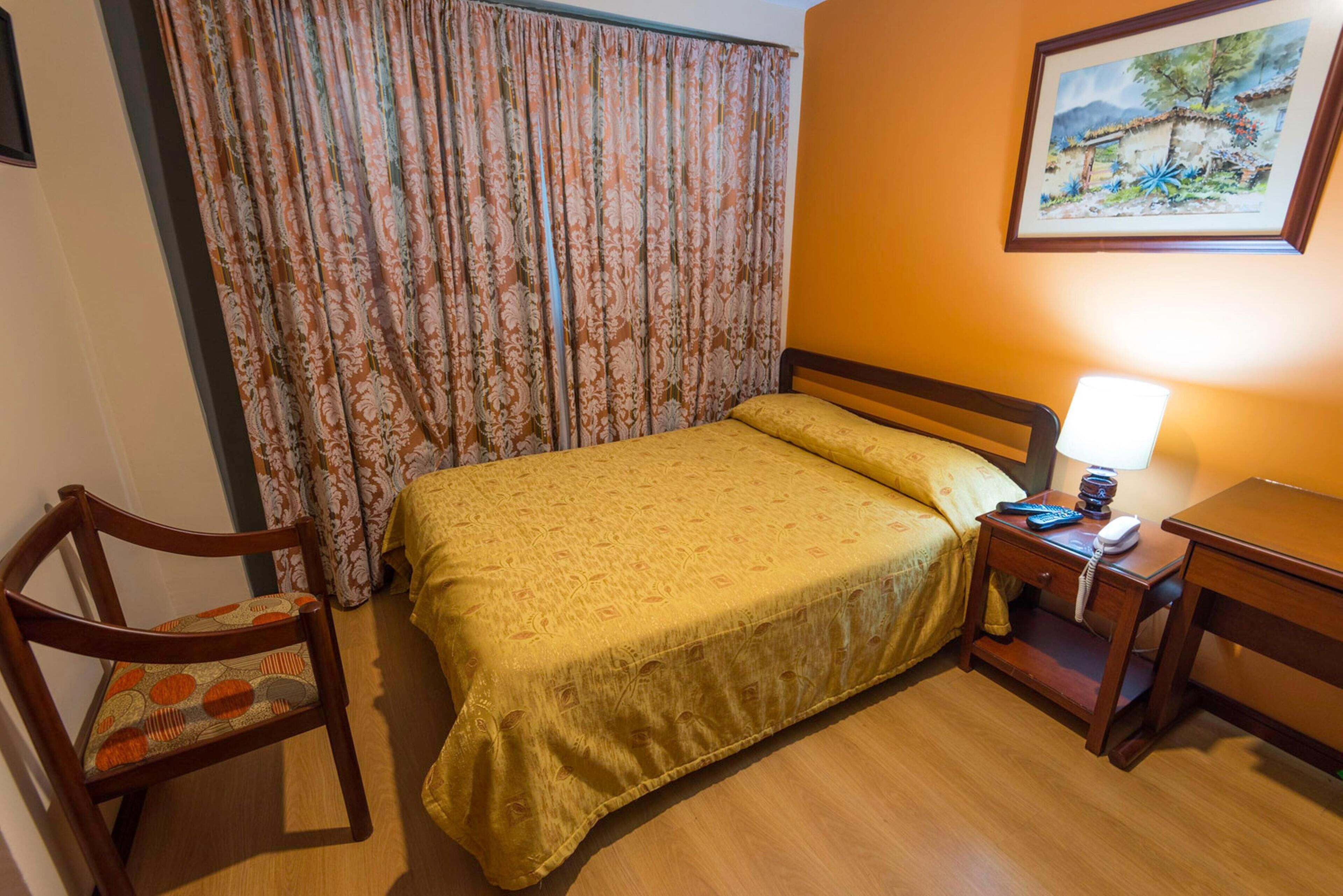 Una habitación de hotel con una cama, una silla, una mesita de noche con lámpara y un cuadro en la pared.