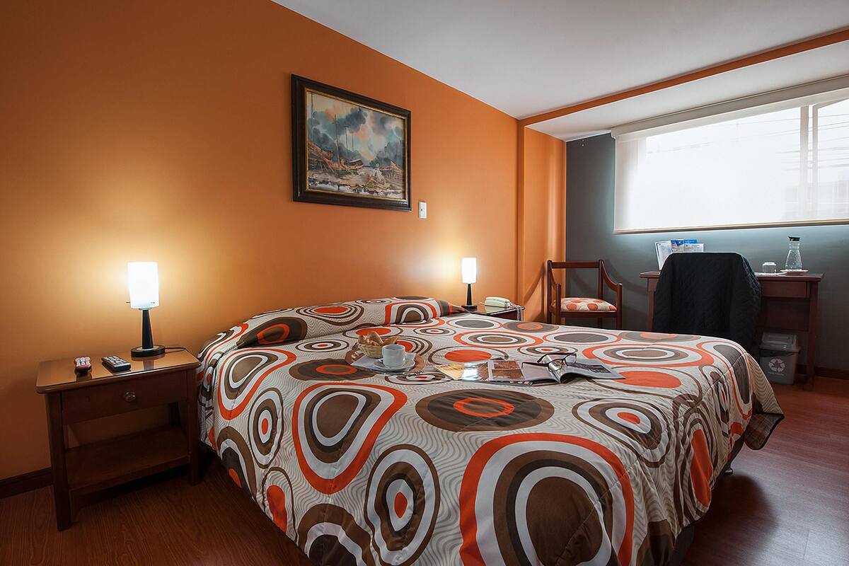 Een hotelkamer met een groot bed, twee nachtkastjes met lampen, een stoel, een bureau met stoel, een raam met jaloezieën en een schilderij aan de muur.